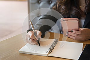 Businesswoman writing on notebook while using smartphone.