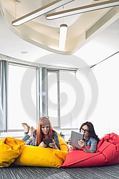 Businesswomen using digital tablets while relaxing on beanbag chairs in creative work space