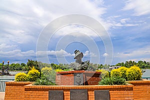 A bust statue of a Spanish explorer on the Savannah riverfront surrounded by red brick and lush green plants and yellow flowers