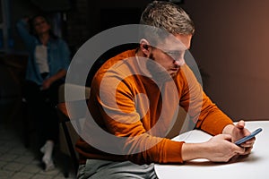 Busy addicted young man using mobile phone sitting at table in dark living room on background of girlfriend sitting on