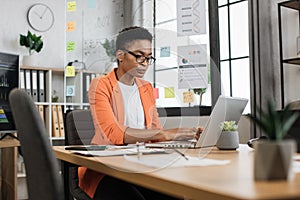 Busy african woman in orange suit sitting at office and using wireless laptop.