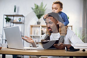 Busy afro man working on laptop with son on neck