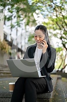 A busy Asian businesswoman is having a business phone call while working on her laptop on the stairs