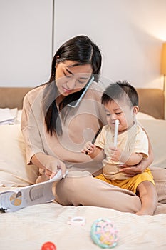 A busy Asian mom is holding her baby boy while talking on the phone and working on documents in bed