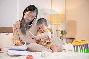 A busy Asian mom is holding her baby boy while talking on the phone and working on documents in bed