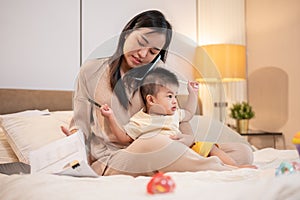 A busy Asian mom is holding her baby boy while talking on the phone and working on documents in bed