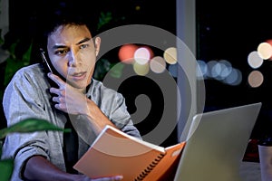 Busy Asian young staff officer man holding a notebook while talking on phone with customer at working desk in office at night,