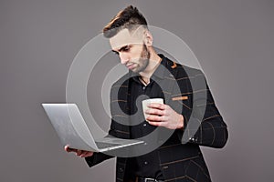 Busy bearded man in business clothes talking by smartphone and using laptop computer while holding cup of coffee in hand