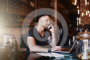 Busy behind the bar. Shot of a young man using a phone and laptop while working in a coffee shop.