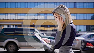 A busy business lady talks on her mobile phone, holding important contracts, she walks by the parking lot where cars are