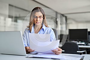 Busy business woman of middle age working on laptop checking documents.