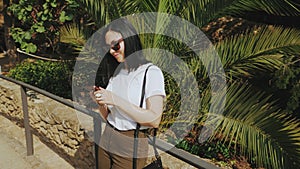 Busy business woman in sunglassess using mobile phone near exotic palm trees. Beautiful young tourist girl surfing