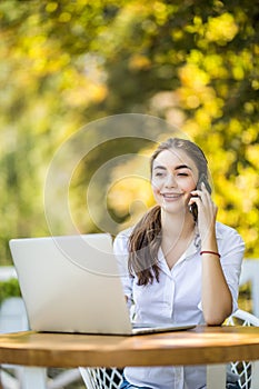 Busy businesswoman talking on mobile phone sitting at the cafe table with laptop Outdoors