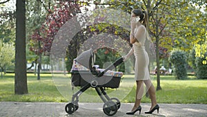 Busy Caucasian businesswoman talking on the phone and rocking baby stroller. Wide shot side view of elegant slim woman