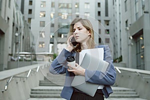 Busy caucasian woman having phone business discussion while holding coffee cup, papers and laptop near office building. Office