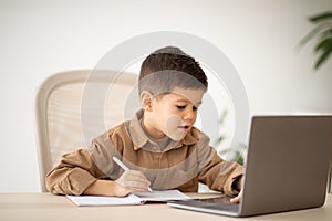 Busy cheerful european small kid has video call with teacher, tutor at computer at table in elementary school