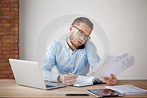 Busy concentrated unshaved businessman in glasses and shirt sitting in a comfortable light office, looking through