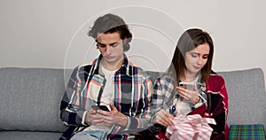 Busy couple sitting on sofa, using smartphones and exchanging gifts