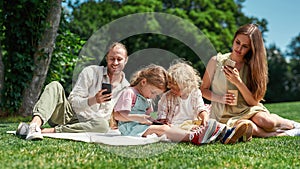 Busy family spending time together outdoors, using smartphone while having picnic in nature on a summer day