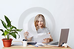 Busy female enterpreneur looking through documents, sitting at table in her cabinet, working with laptop computer, smart phone and