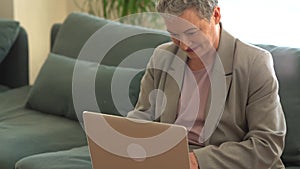 Busy gray-haired lady in a business suit is working with a laptop at home alone while sitting on the couch. Remote