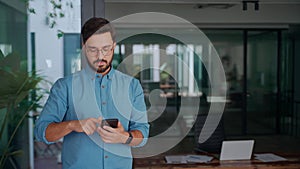 Busy happy young latin business man holding smartphone standing in office.