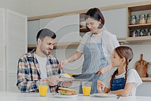Busy housewife holds frying pan, gives prepared meal to husband and daughter, have delicious breakfast together, sit at kitchen