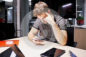 Busy male entrepreneur in casual t shirt sitting at table and working on project online while browsing cellphone