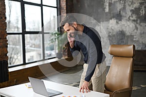 Busy male office employee standing near office desk, talking on the smartphone