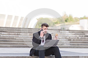 A busy man in a business suit sits on laptops with a laptop on his lap, eats a sandwich and speaks on a mobile phone