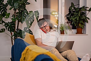 Busy man sitting on couch with laptop. Serious guy with glasses talking on phone, focused on work