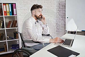Busy man sitting on a wheelchair, talking on a phone and working on a laptop