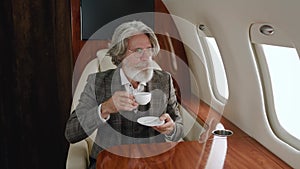 Busy mature business man looking on his watch and then at window while travels on private plane. Rich senior man