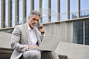 Busy middle aged business man making phone call using laptop sitting outside.
