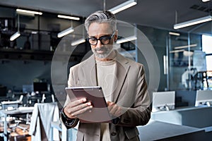 Busy middle aged business man wearing suit standing in office using tablet.