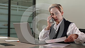 Busy middle-aged business woman in suit working at office, sitting on work desk with laptop, holding documents, having