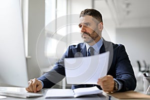 Busy middle aged male entrepreneur working with computer and documents, checking data and financial reports in office