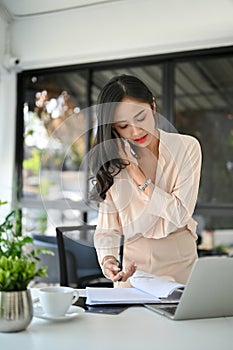 Busy millennial Asian businesswoman is on the phone with a business client, standing at her desk