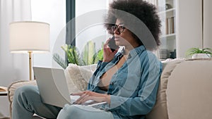 Busy multitasking African American girl business woman talking mobile phone work laptop computer online at home couch