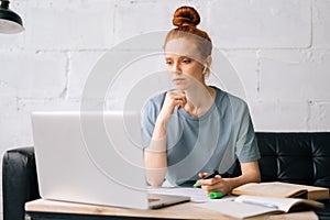 Busy pretty redhead young woman student is taking notes in workbook by looking at laptop screen.