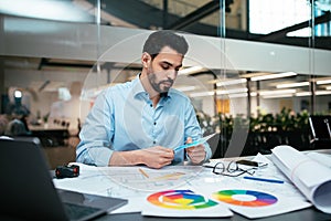 Busy serious attractive millennial arab guy designer with beard works with color palettes at workplace