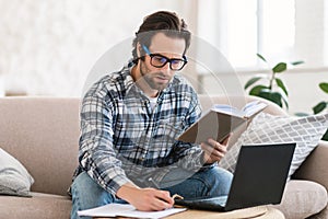 Busy smart millennial european male student in domestic clothes with stubble in glasses with book and pc
