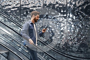 Busy, smiling handsome man using mobile phone, while going down on escalator of shopping mall.
