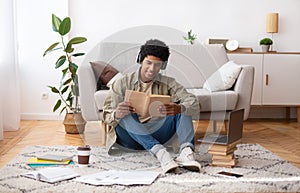 Busy teenage male student in headphones reading book during online class near laptop computer indoors