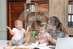 Busy woman trying to work while babysitting two daughte.  Young beautiful Asian mother holding little baby on lap while another