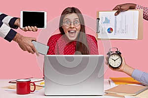 Busy woman wears spectacles, being overworked, has deadline for preparing task, surrounded with laptop, hands hold
