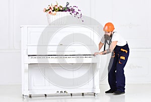 Busy worker in orange protective helmet and blue overall moving grand piano isolated on white background. White piano