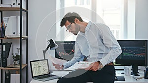 Busy working day. Young successful trader or businessman in formal wear and eyeglasses sitting at modern office in front