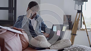 Busy young African American man receiving phone call sitting on couch in home office with laptop. Portrait of confident