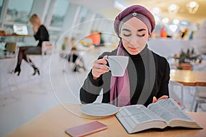 Busy young arabain woman read book and drink coffee. She it turning picture. She sits in side. Woman is busy.